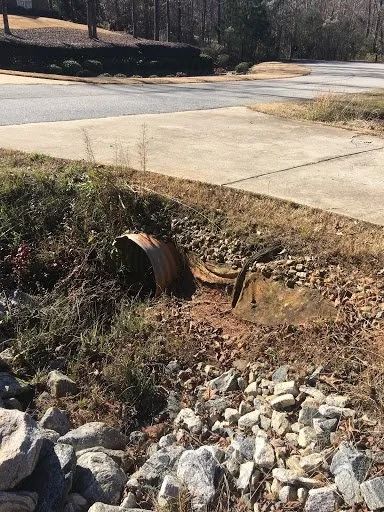 A rusted corrugated metal drainage pipe sits partially buried in an overgrown ditch beside a paved driveway.