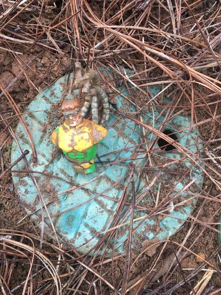 A plastic superhero figurine standing on a circular, teal utility cover nestled among pine needles and dirt.