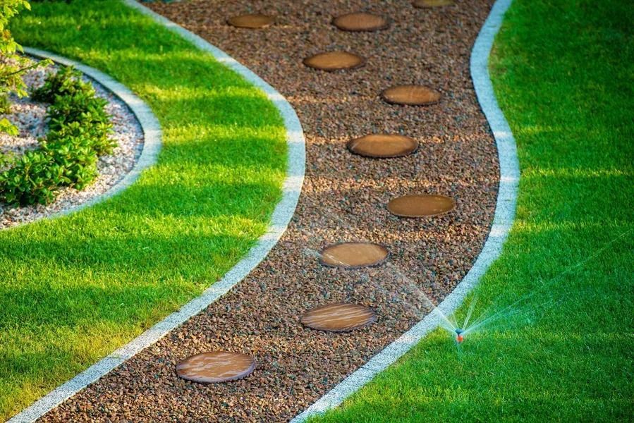 A winding gravel garden path with round stepping stones, flanked by vibrant green lawn and a sprinkler.
