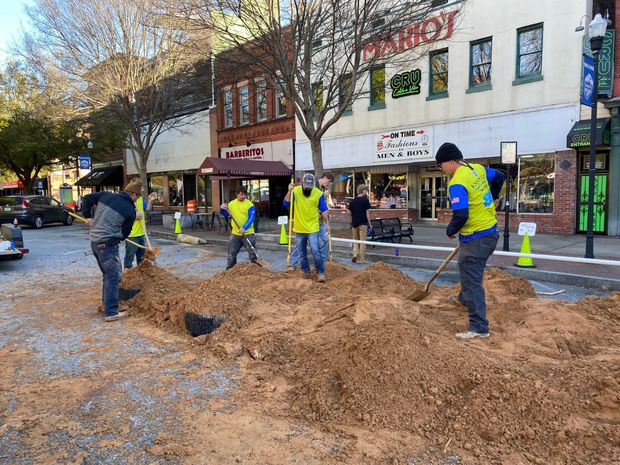 Workers in neon vests dig in a dirt-filled street in front of downtown storefronts.