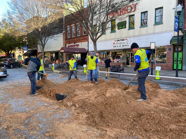 Workers in neon vests dig in a dirt-filled street in front of downtown storefronts.