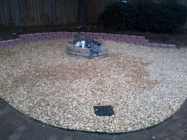 A gravel patio features a circular stone fire pit and a black drain, enclosed by a low red brick retaining wall.