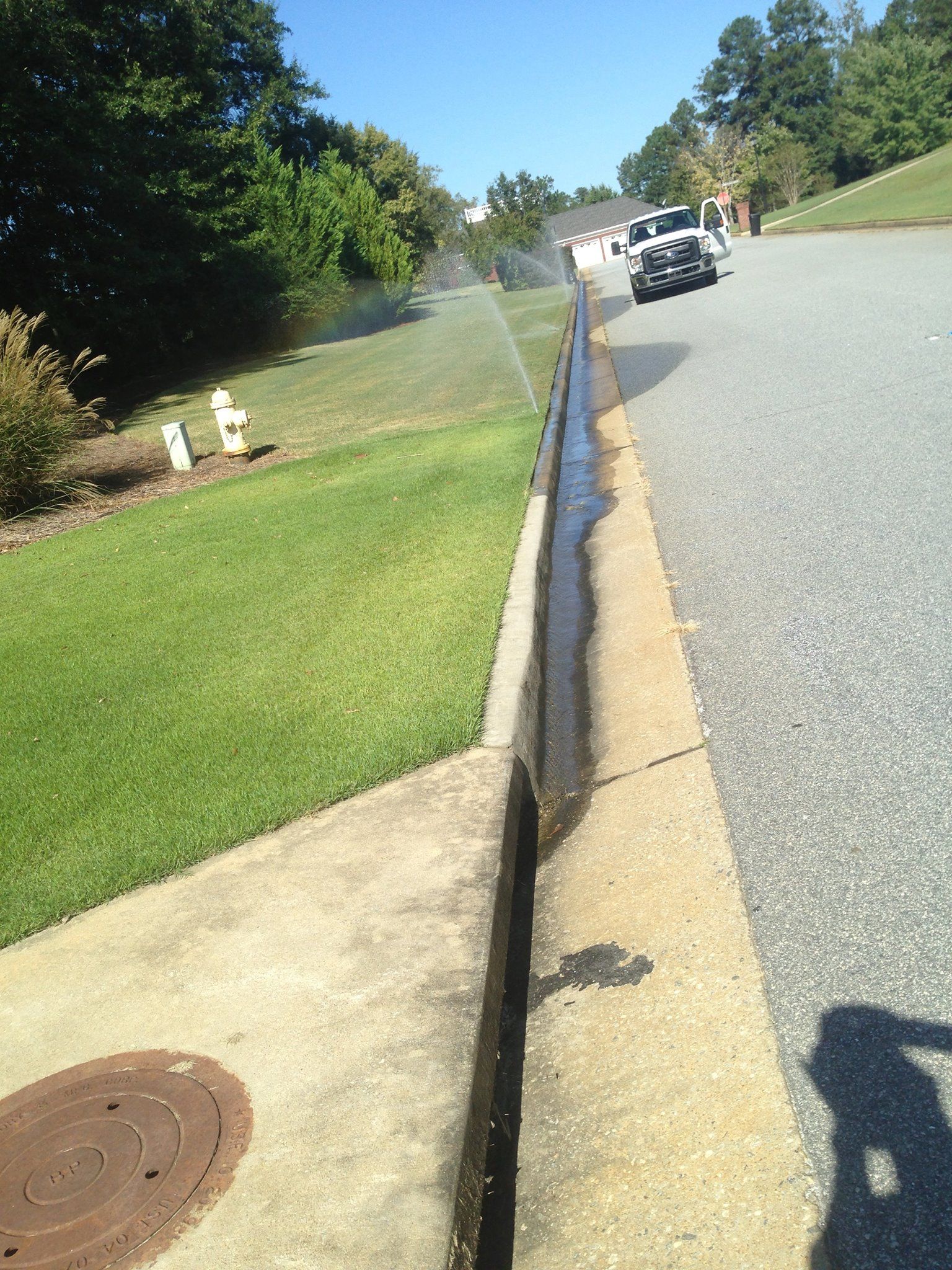 A roadside sprinkler system creates water runoff that flows into a concrete gutter along an asphalt street.