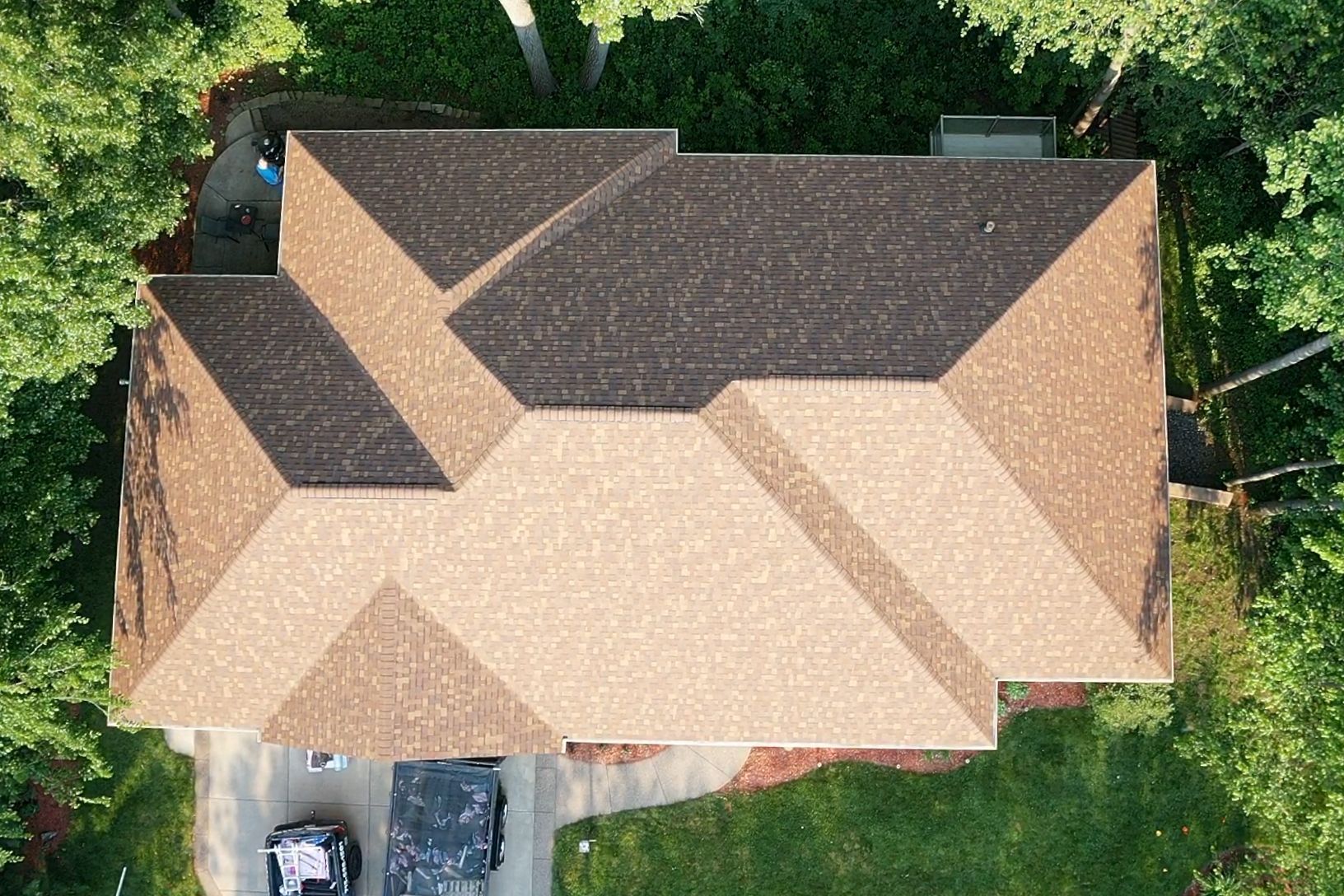Aerial view of a tan shingled house roof surrounded by green trees, with a driveway and parked car visible.
