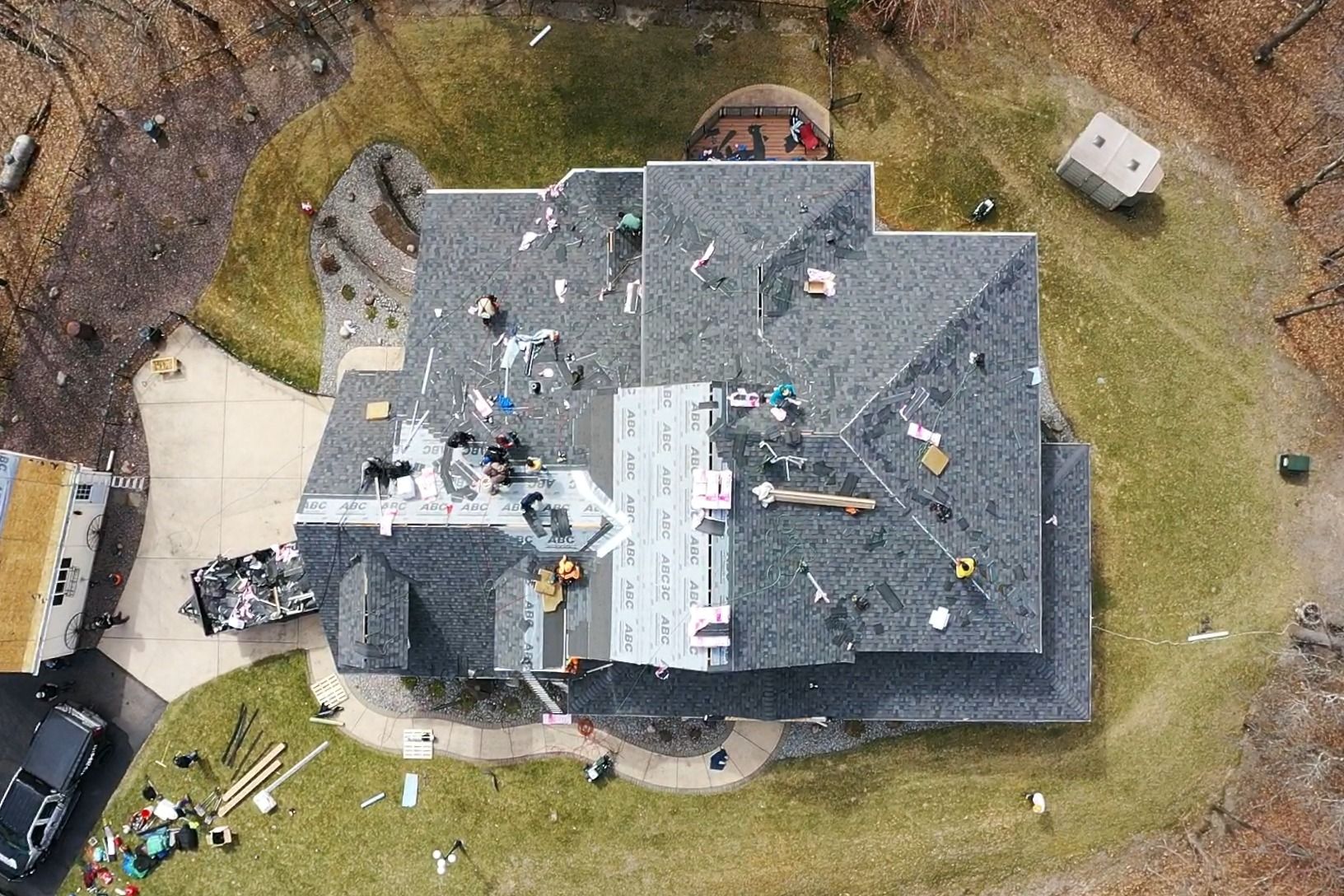 Aerial view of workers repairing a gray roof on a house with a yard and nearby equipment