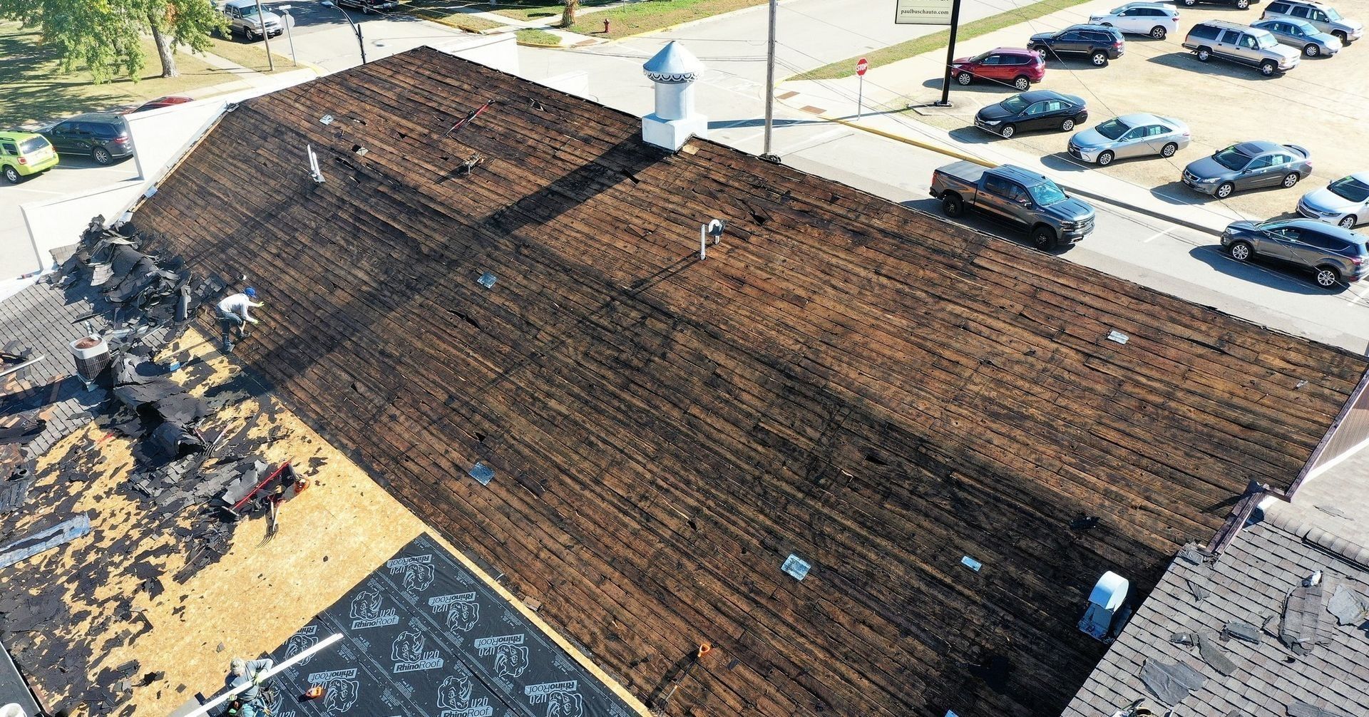 Aerial view of a large brown shingled roof with skylights beside a parking lot and trees