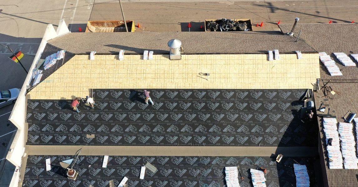Rooftop under construction with plywood sheathing, roofing materials, and workers near the edge
