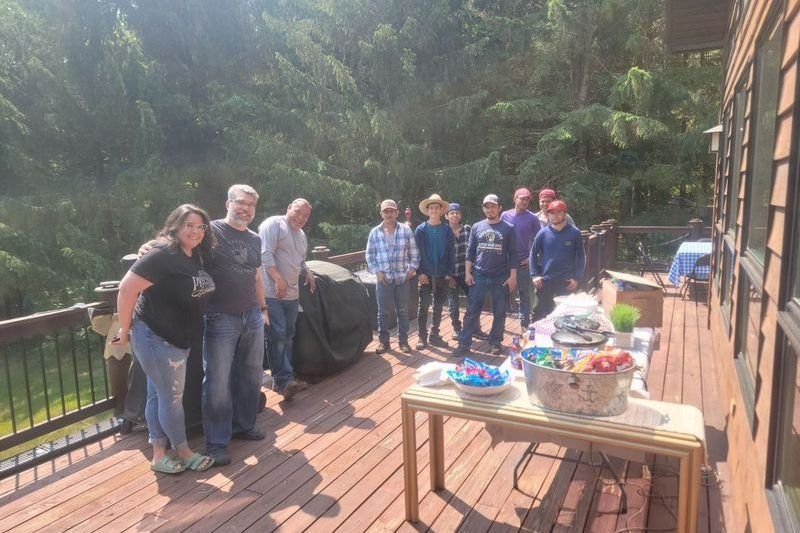 Group gathered on a sunlit wooden deck beside a table with food and drinks, near a forested backdrop.