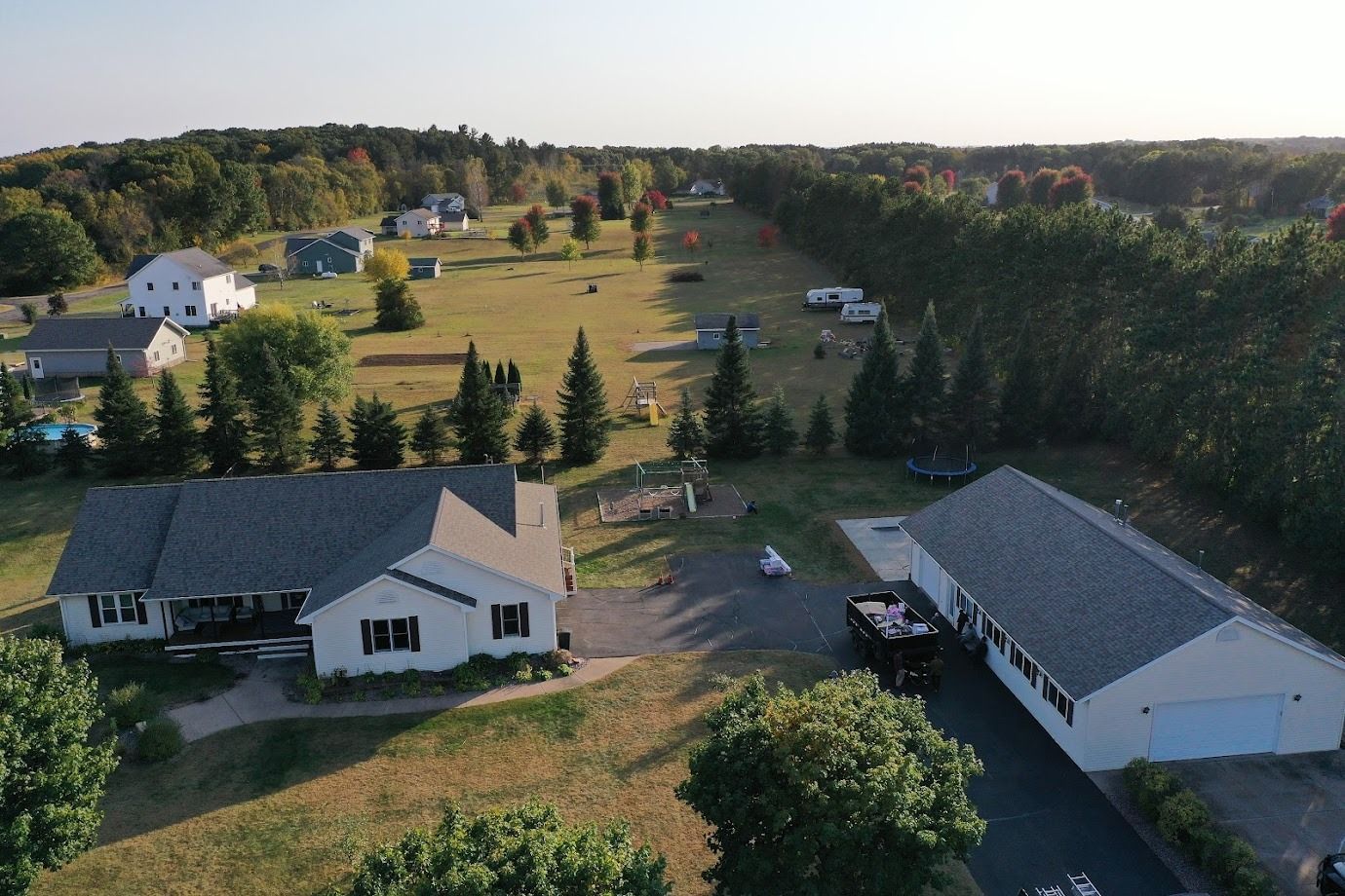 Aerial view of a rural neighborhood with houses, a driveway, and fields bordered by trees at sunset.