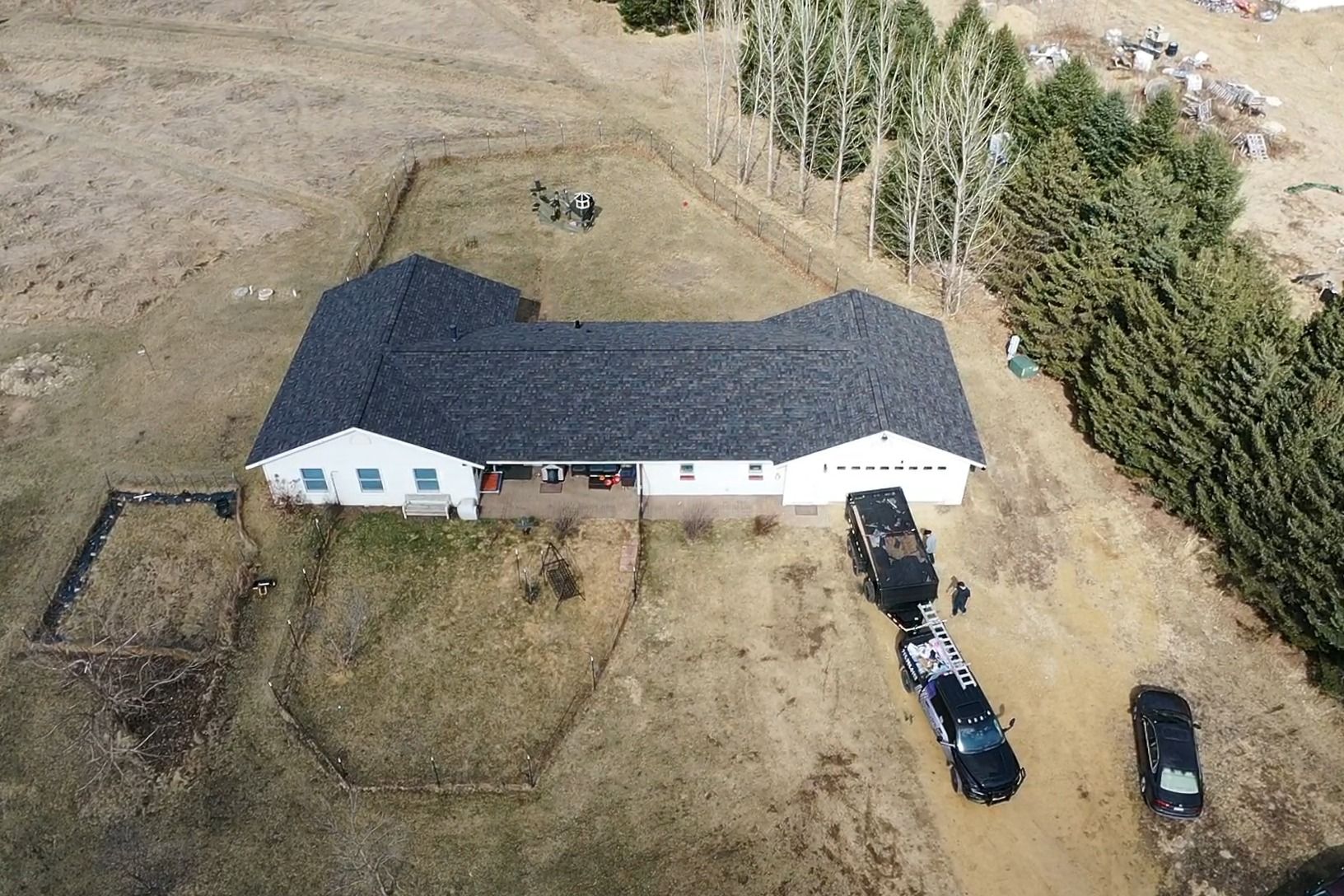 Aerial view of a white house with a dark roof, parked vehicles, and muddy ground in a rural area