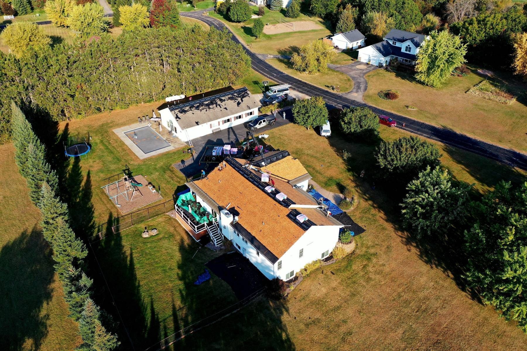 Aerial view of a rural house with a red roof, yard, and surrounding trees along a road.