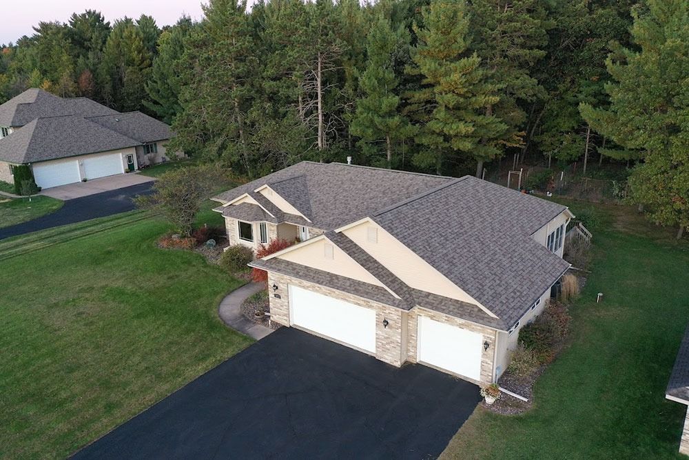 Aerial view of a beige suburban house with a two-car garage, driveway, and trees behind it.