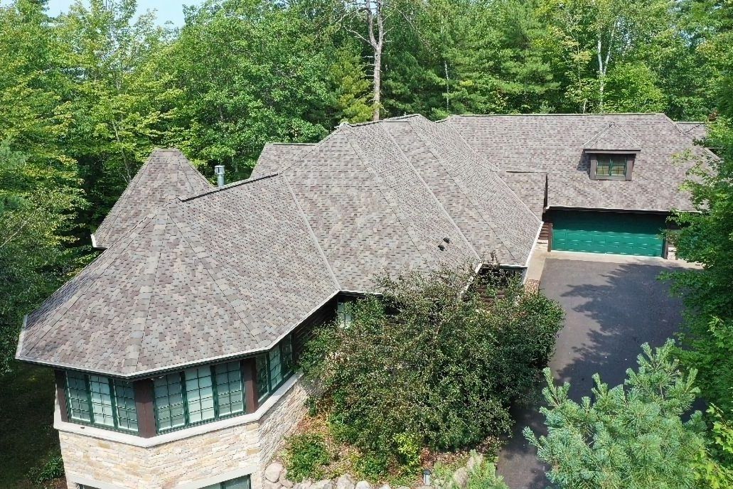 Aerial view of a large shingled house with a driveway, surrounded by trees and greenery