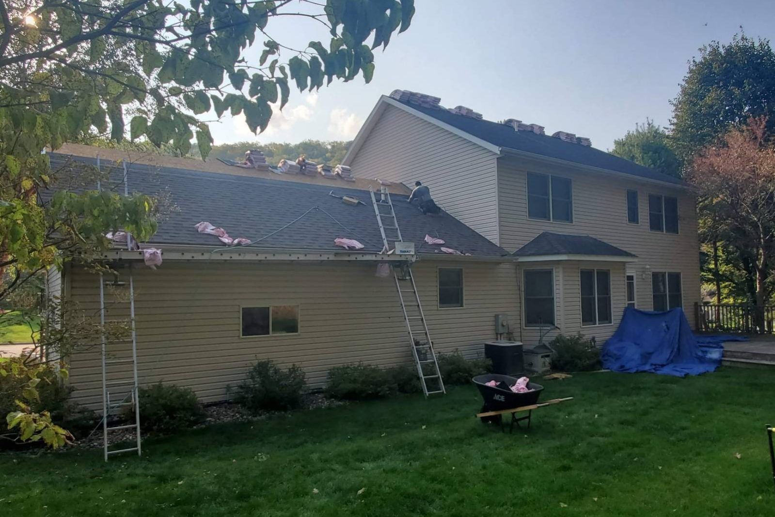 Suburban house with roofers on ladders, blue tarp on side, and a lawn in front