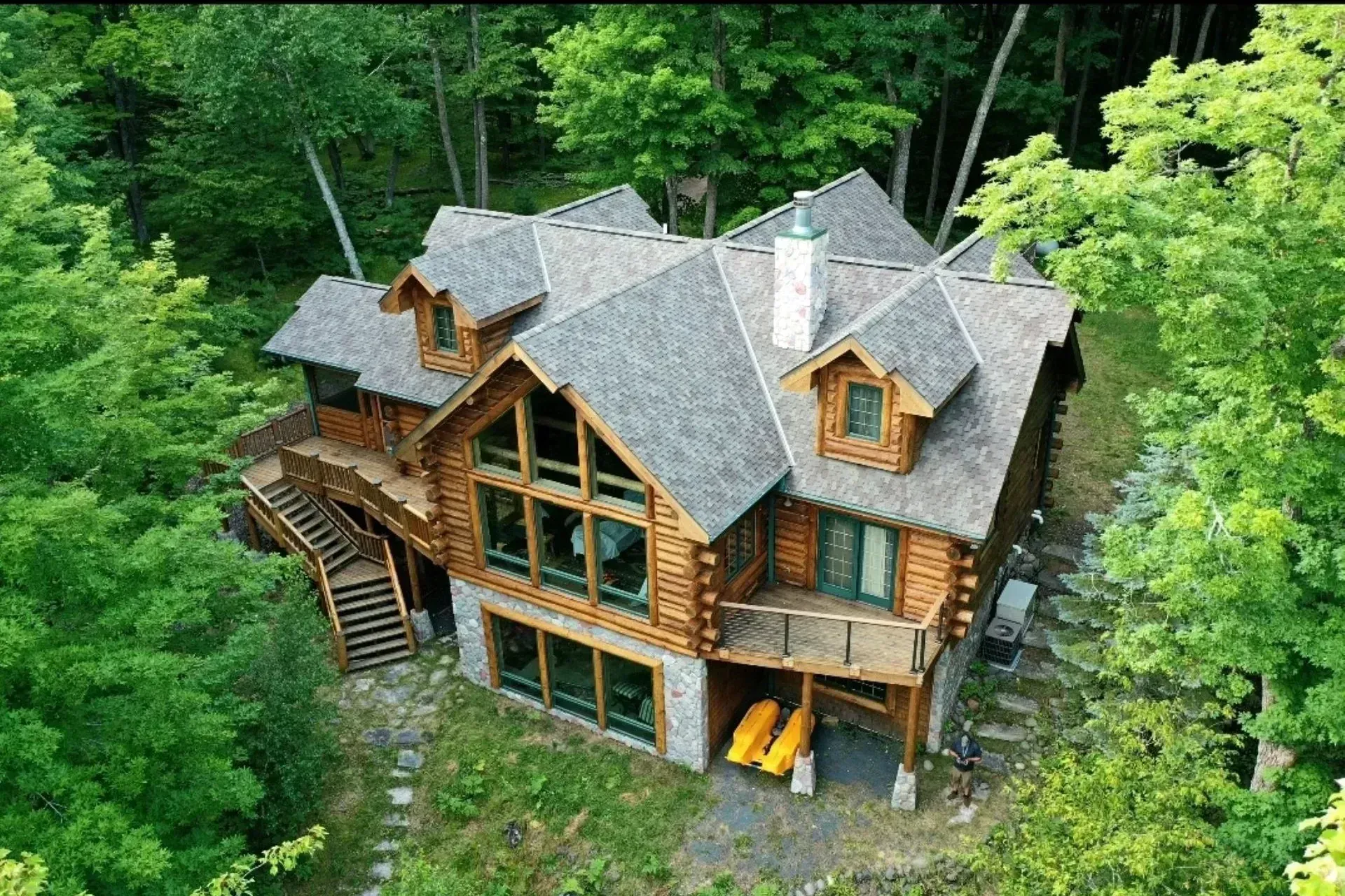 Aerial view of a large rustic log cabin with a gray roof, surrounded by dense green forest.