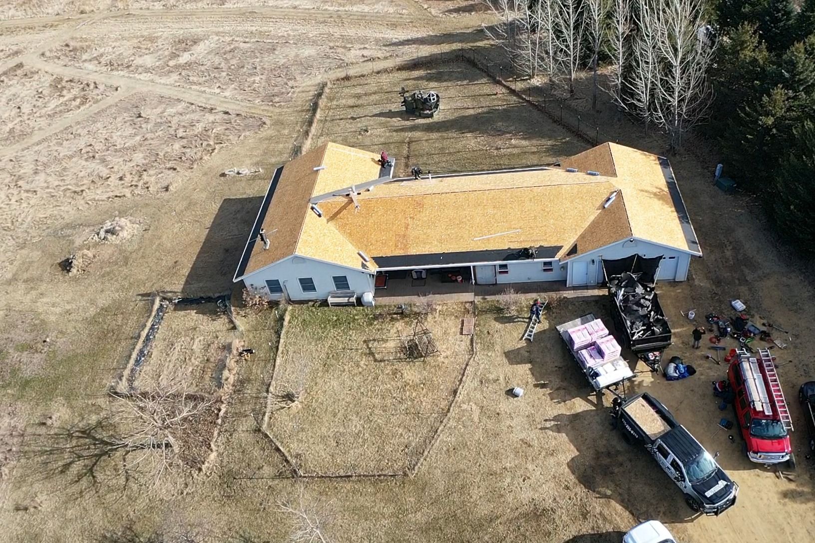 Aerial view of a house with a yellow roof, fenced yard, and several vehicles parked beside it.