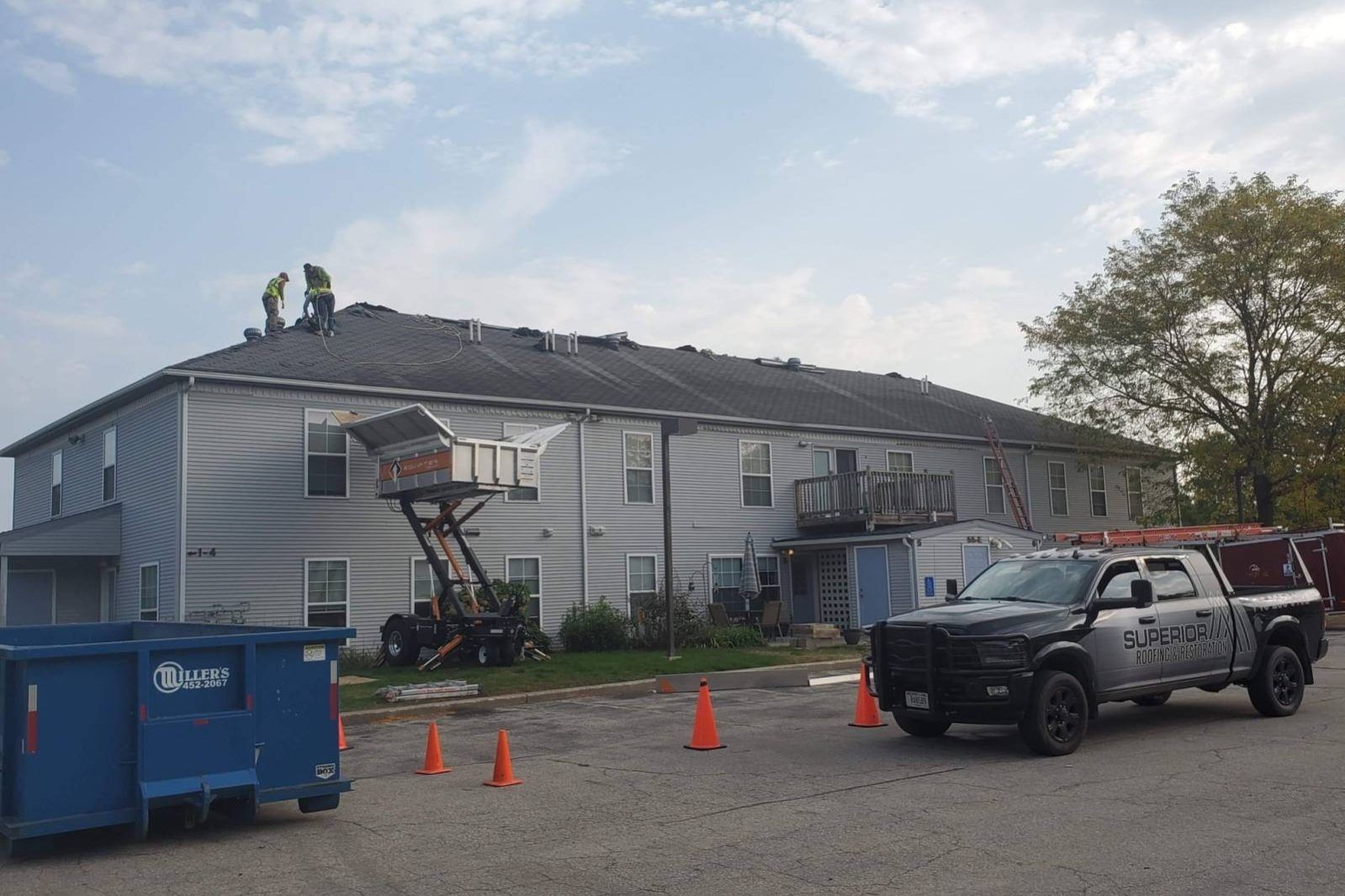 Apartment building with roof damage, a boom lift, and a pickup truck in a gravel lot