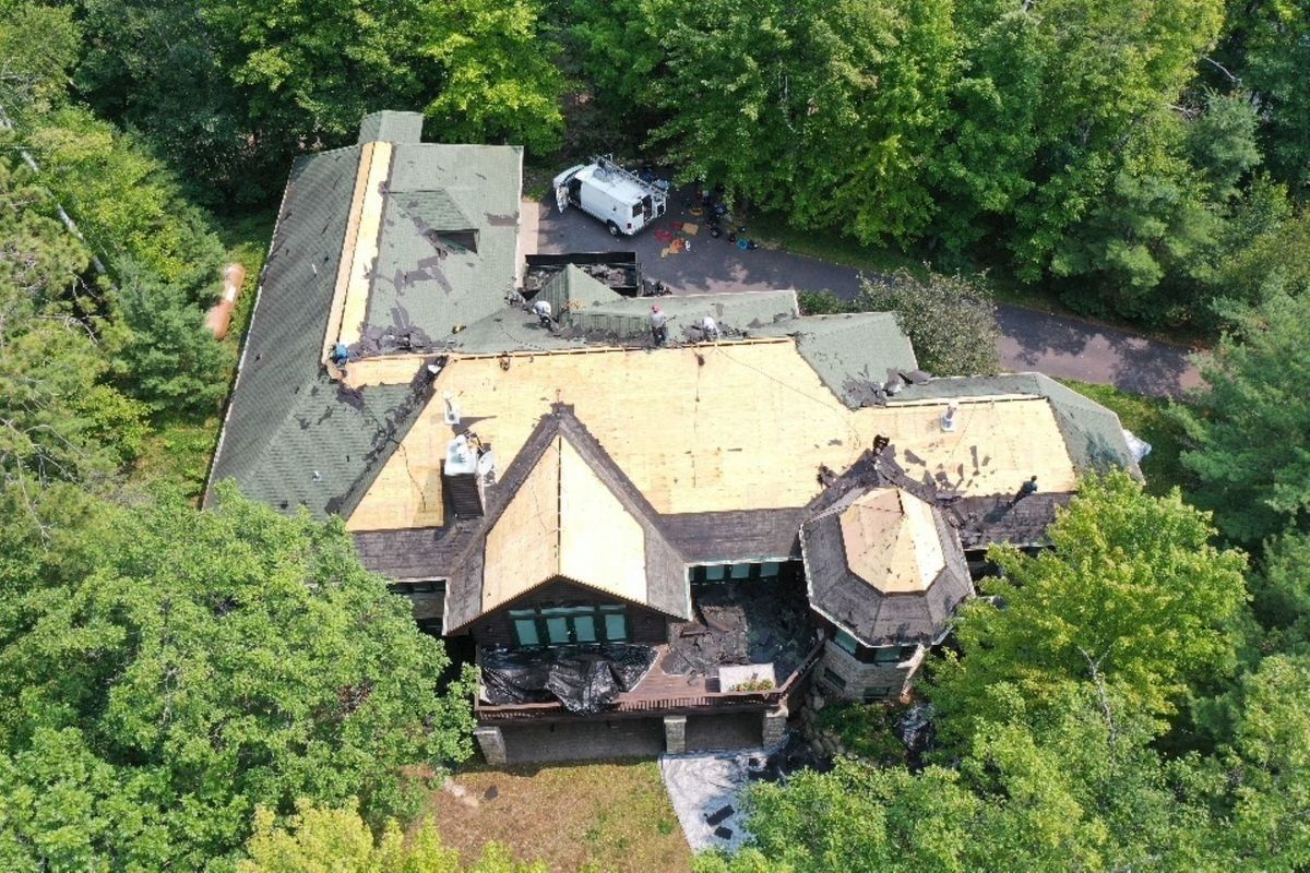Aerial view of a large house with a gray roof surrounded by dense green trees