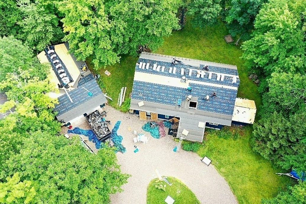 Aerial view of two small cabins in a green wooded clearing with a gravel path and scattered outdoor items.