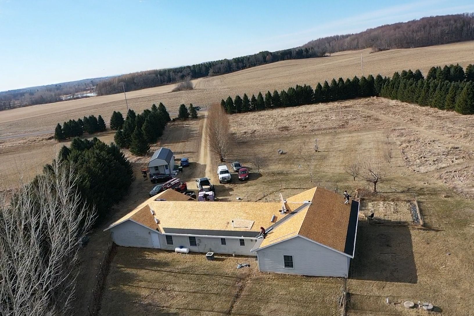 Aerial view of a farmhouse with tan roofs, parked vehicles, and open fields bordered by trees.