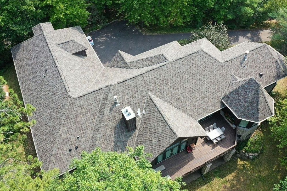 Aerial view of a large house with a gray shingled roof surrounded by dense green trees.
