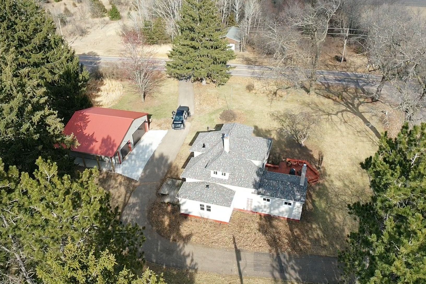 Aerial view of a house with a red-roofed shed, driveway, and trees in a rural yard.