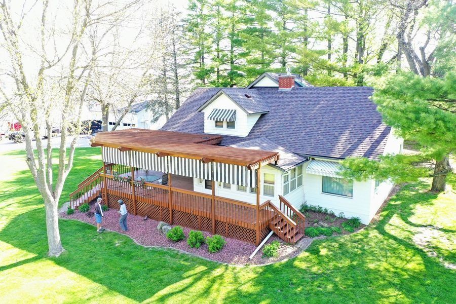 White house with dark roof and wooden porch, surrounded by green lawn and trees.