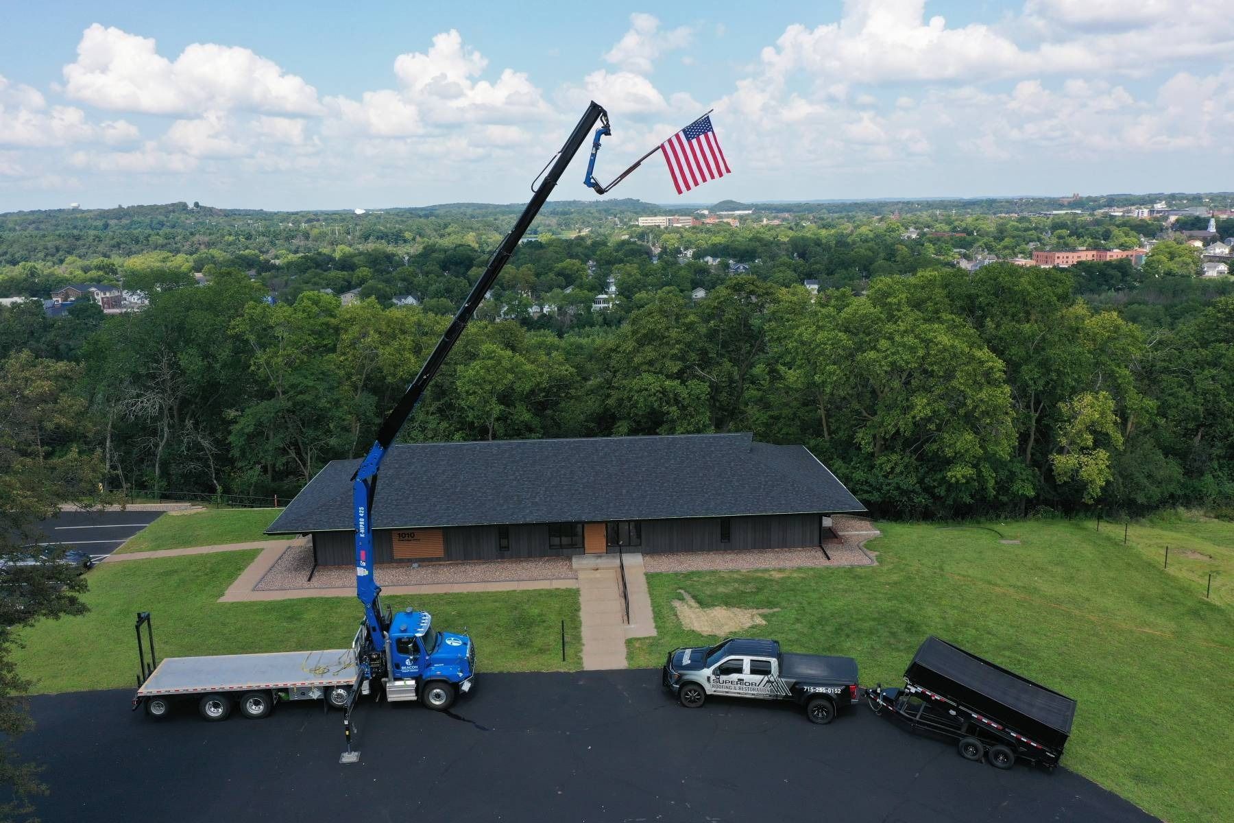 Crane truck lifting an American flag beside a house and vehicles under a bright sky
