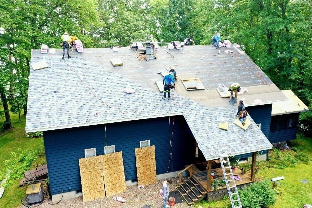 Workers repairing a blue house roof surrounded by trees, with ladders and plywood sheets underway