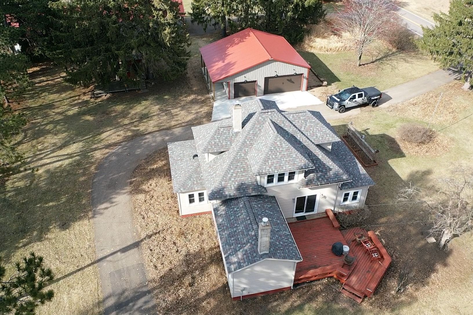 Aerial view of a gray house with a red deck, driveway, and detached garage surrounded by trees.