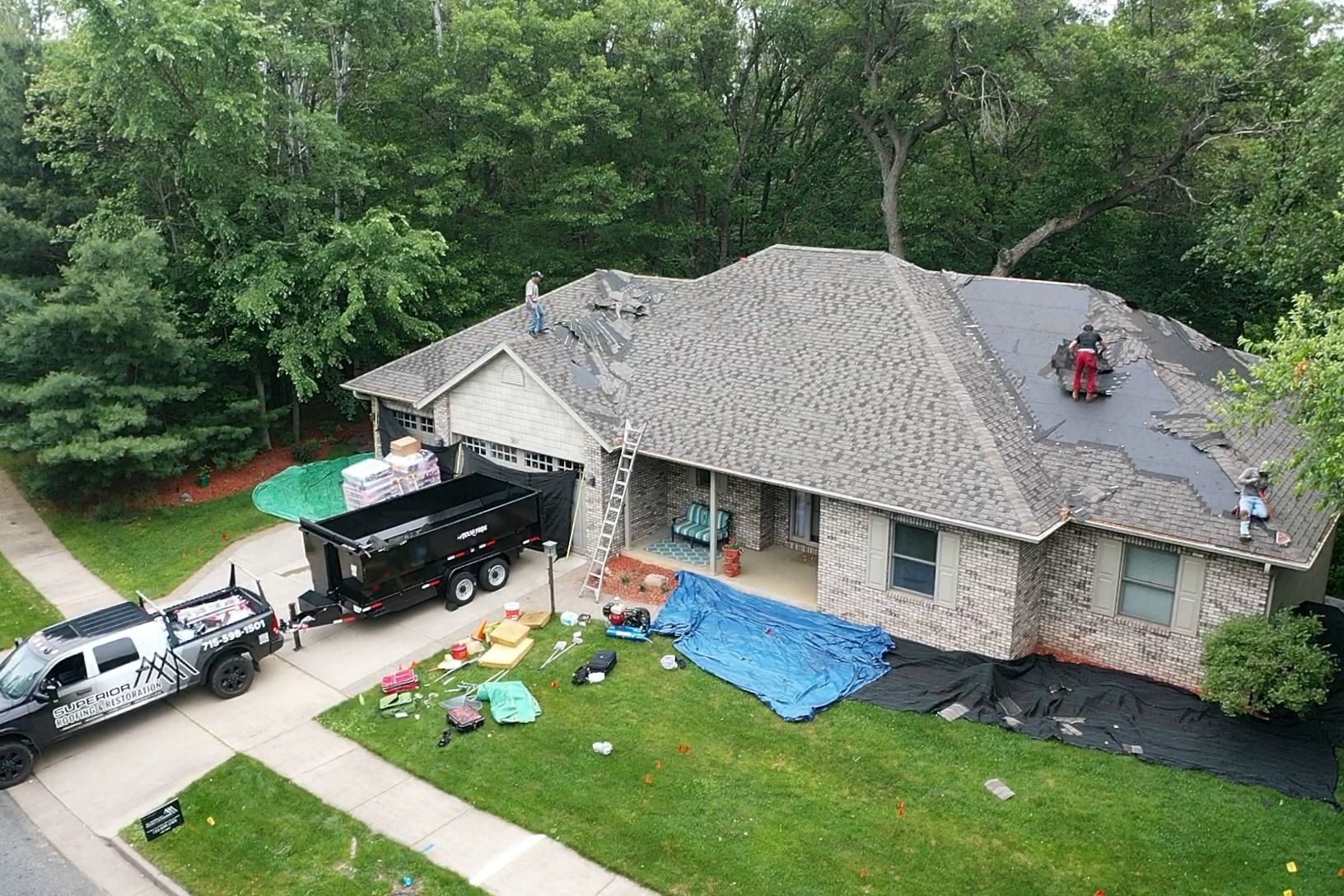 House with roof repair supplies and workers on the lawn beside parked trucks, surrounded by trees