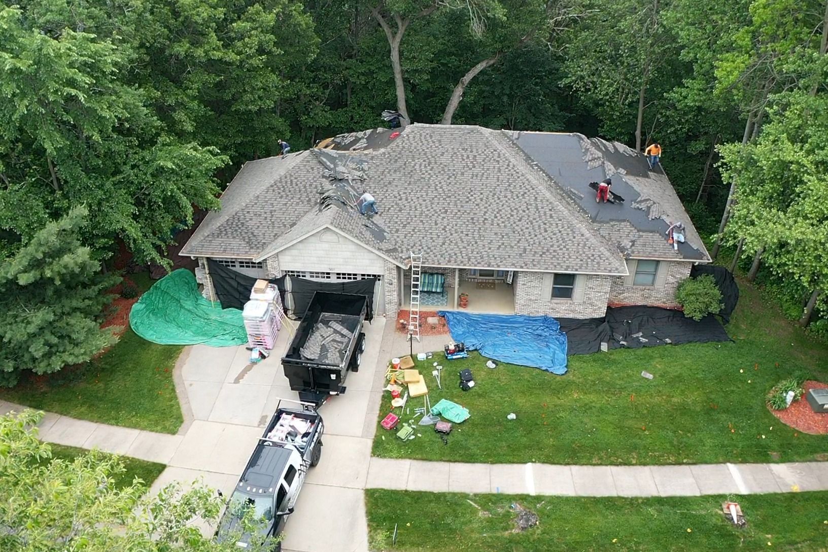 Aerial view of a house with a damaged roof, debris in the driveway, and tarps on the lawn and roof