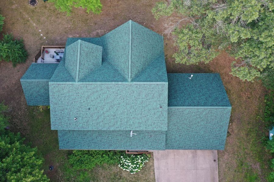 Aerial view of a teal-roofed house with a driveway and surrounding trees.