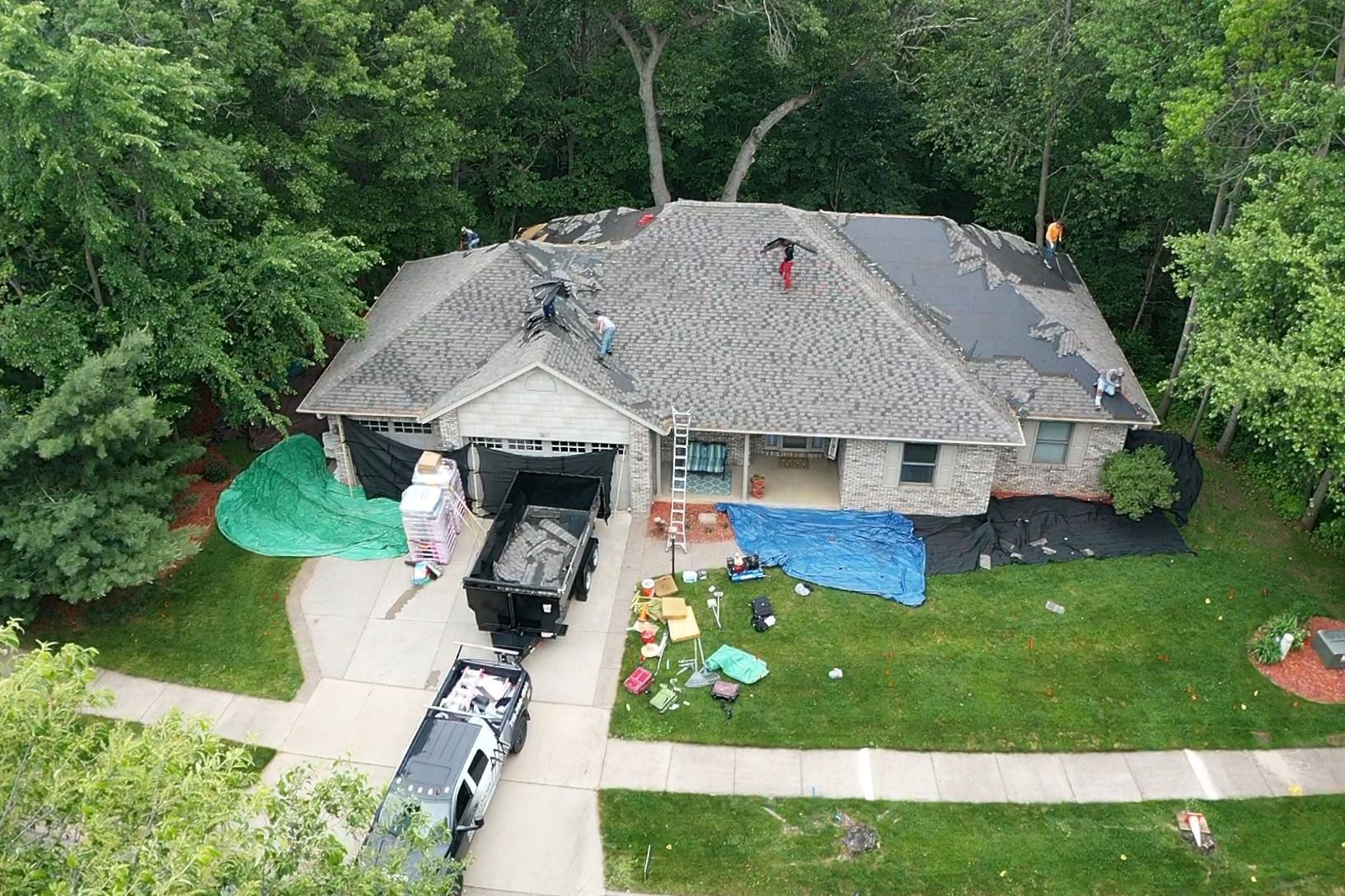 Aerial view of a house with roof damage, debris in the driveway, and tarps on the yard after storm cleanup