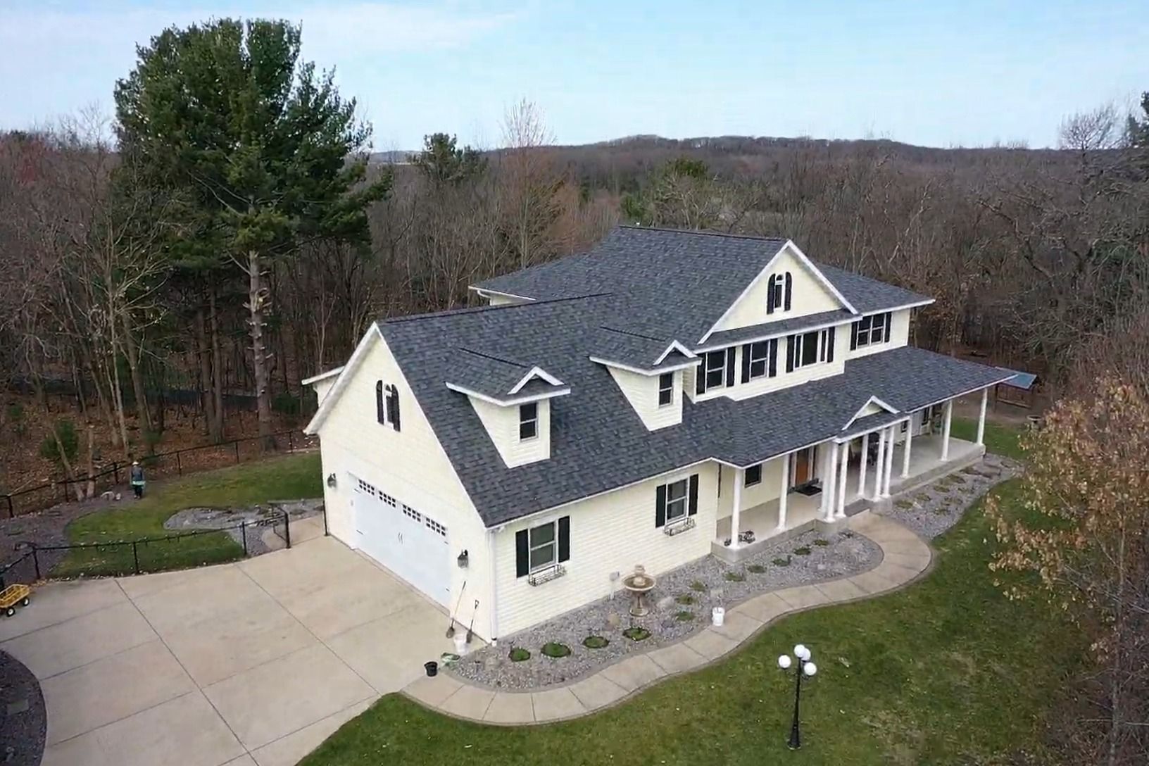 Aerial view of a white suburban house with a dark roof, driveway, porch, and trees surrounding it.