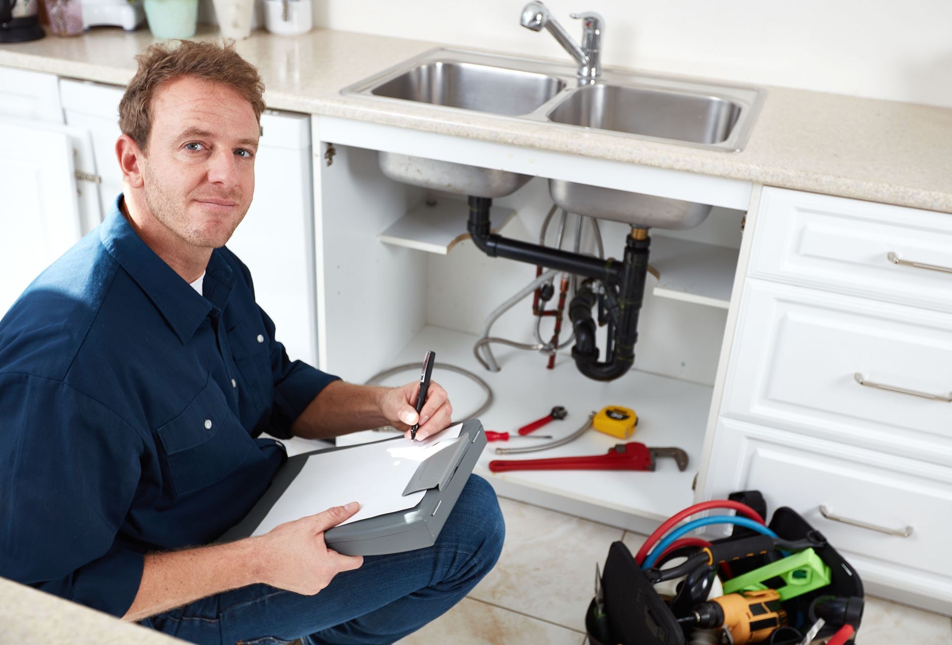 Plumber kneeling under a kitchen sink, writing on a clipboard, tools nearby.