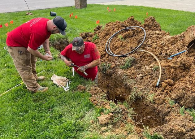 Two workers in red shirts repair underground utility lines in a grassy yard.