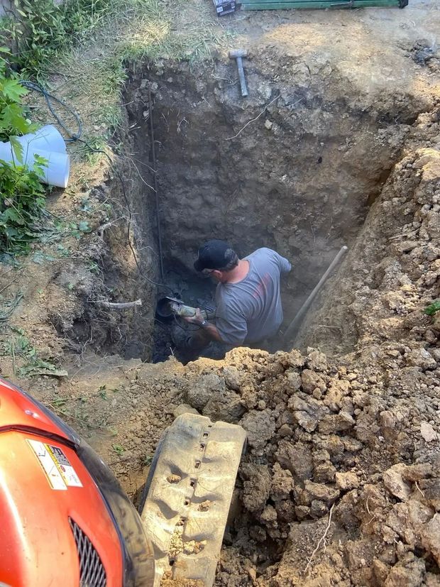 Man digging in a deep, square hole with a shovel. Dirt surrounds him, equipment visible nearby.
