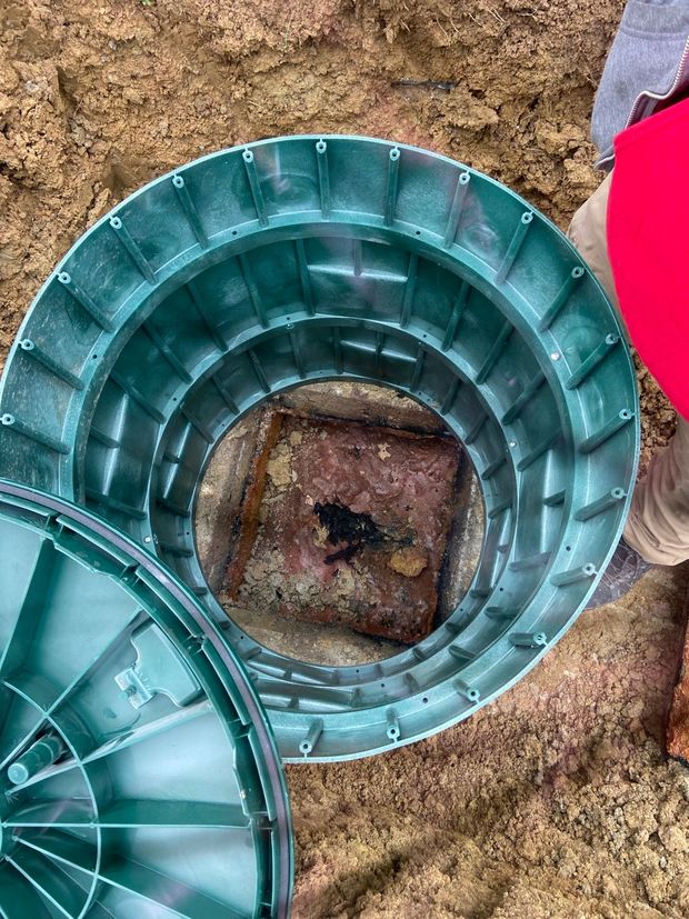 Green septic tank lid open in the dirt, revealing a square opening.