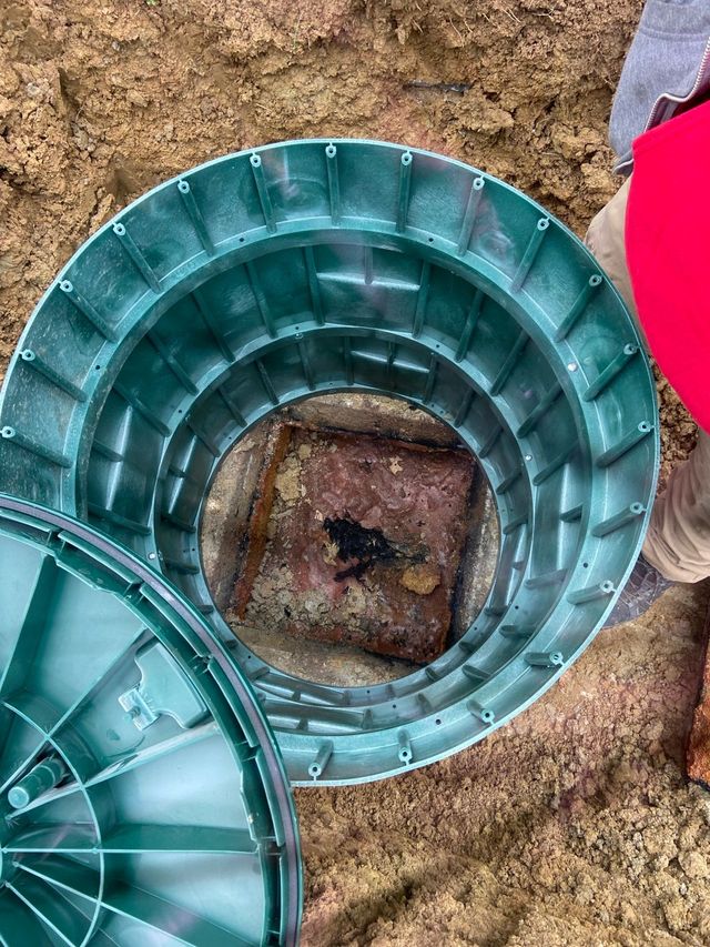 Green septic tank lid open in the dirt, revealing a square opening.