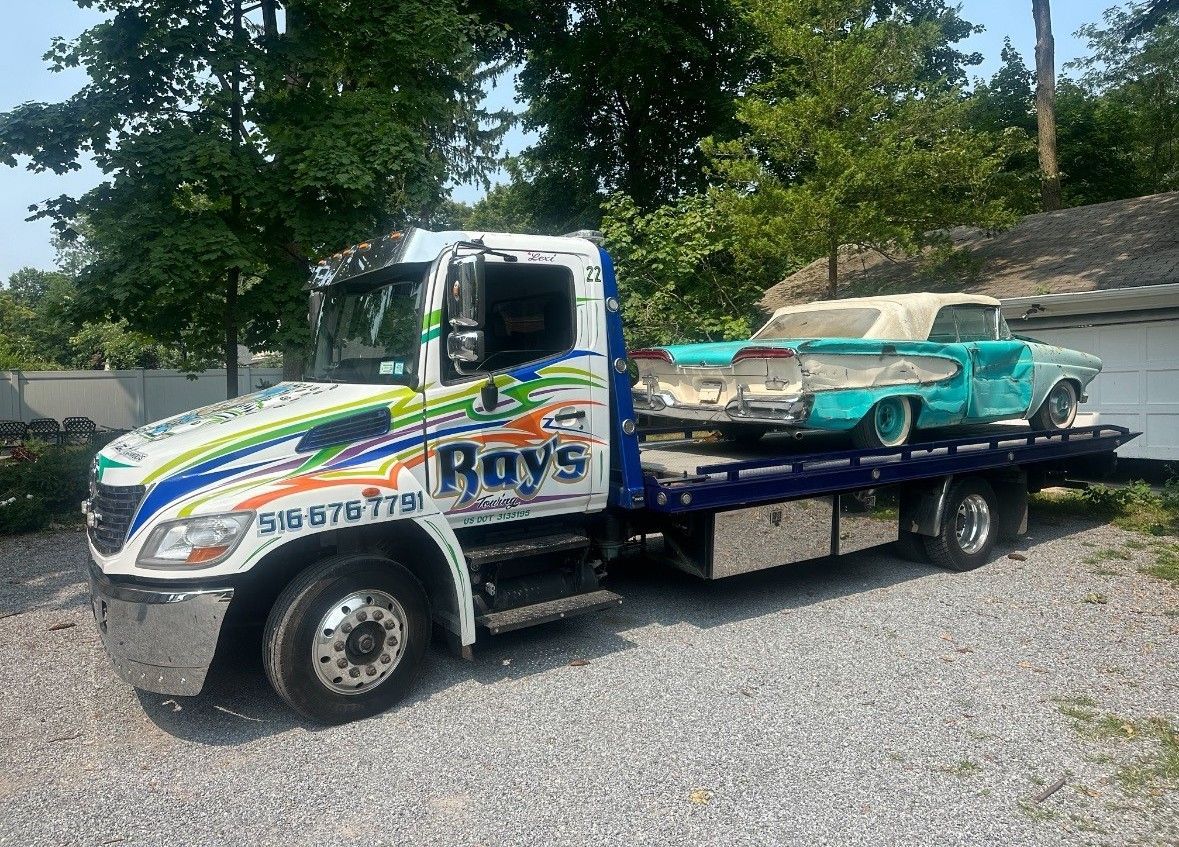 A tow truck with two cars on the back is parked in a gravel lot.