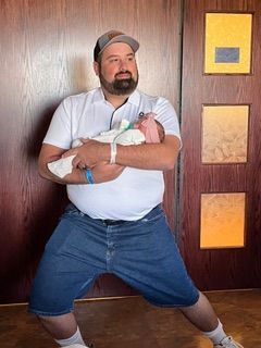 Man in white shirt and denim shorts holding a newborn baby, posing by a wood-paneled wall.