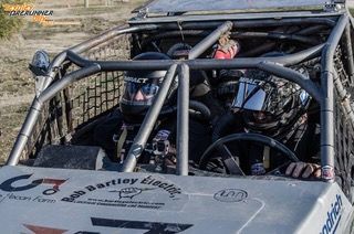 Two people in race car, wearing helmets, driving on dirt track.