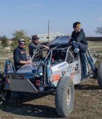 Three people pose with a silver off-road buggy outdoors on a sunny day.