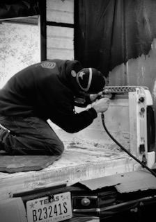 Man kneeling, working on truck bed with tool, wearing a cap.