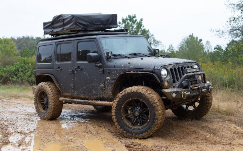 A black jeep with a tent on top of it is driving through a muddy field.