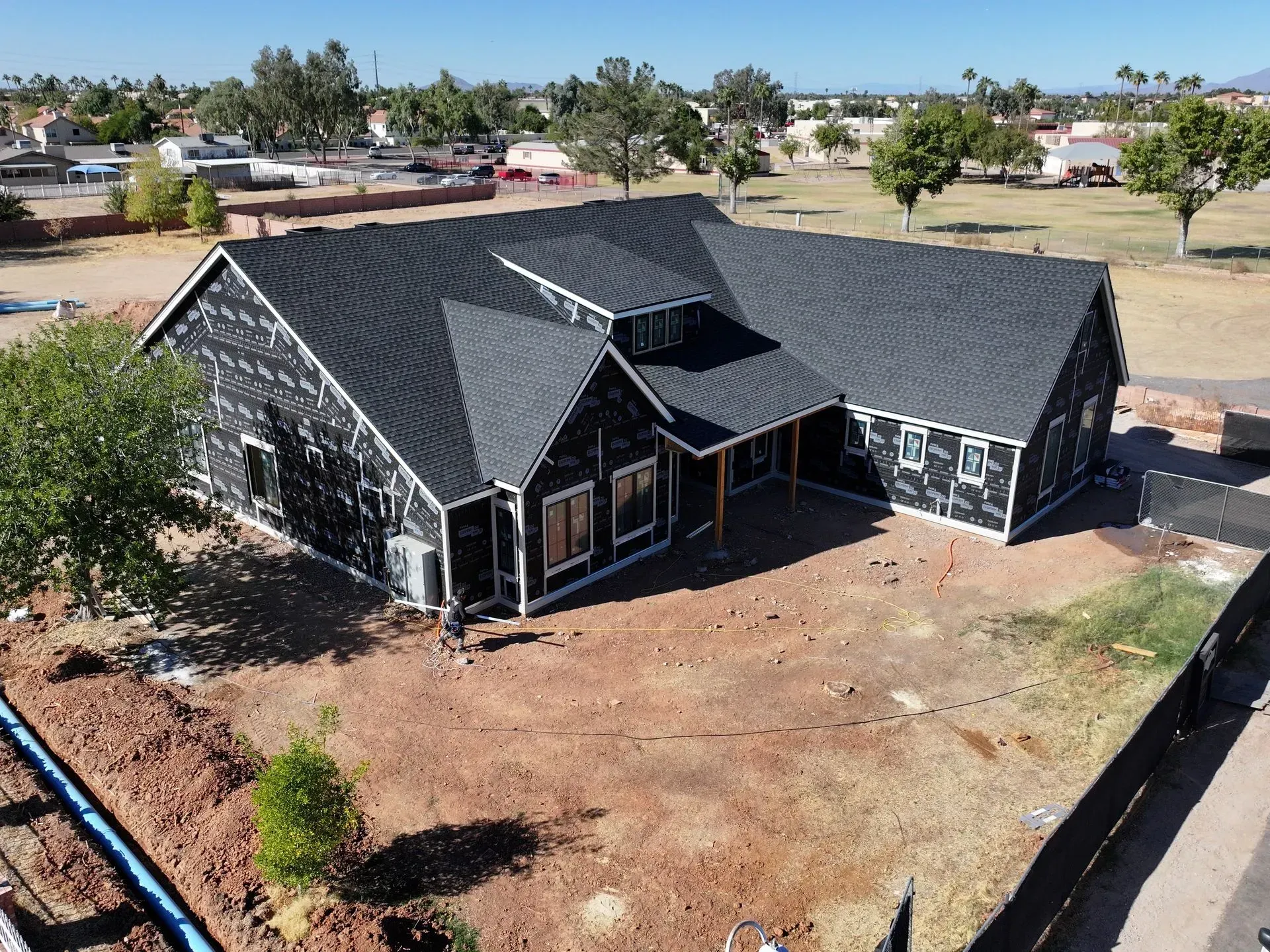 Aerial view of a dark-roofed house with a large dirt yard and nearby trees in a suburban area