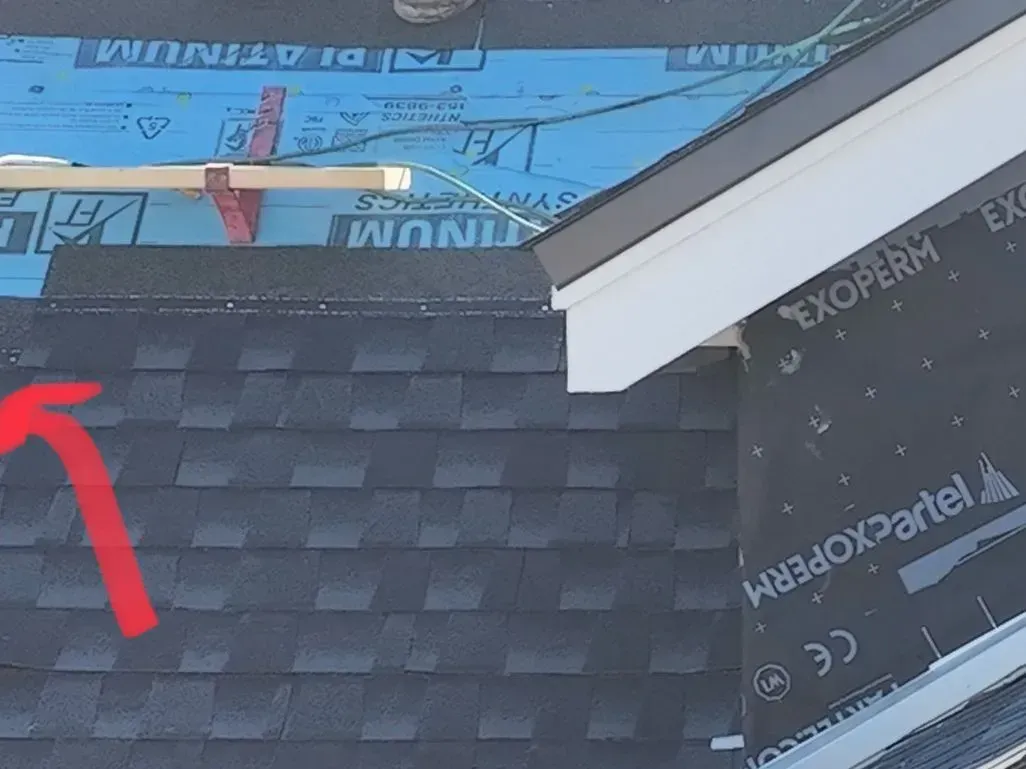 Roof shingles with red arrow pointing to the edge near a white roof trim and house wrap below