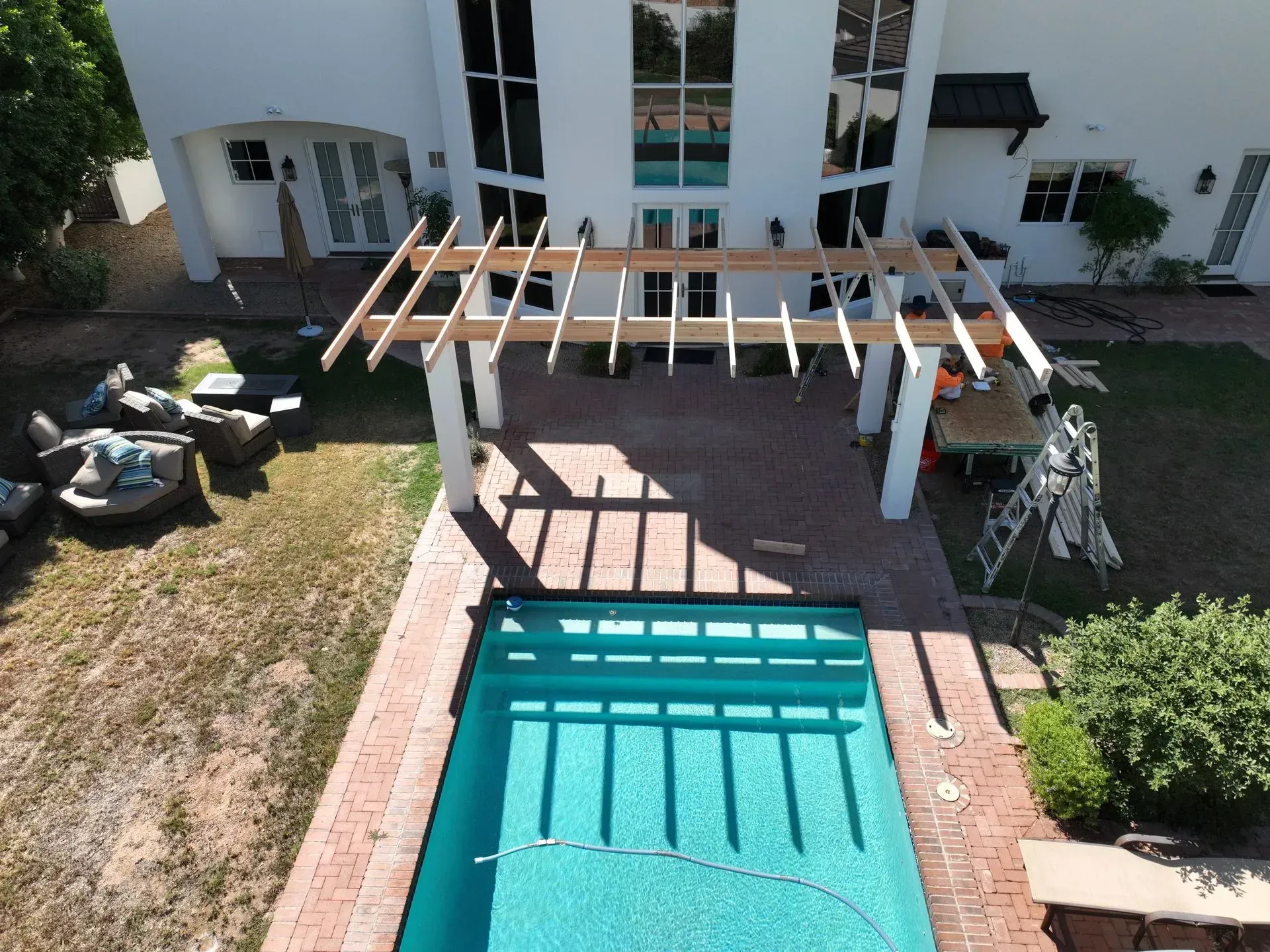 Backyard pool beside a white house with a pergola and patio seating, viewed from above.