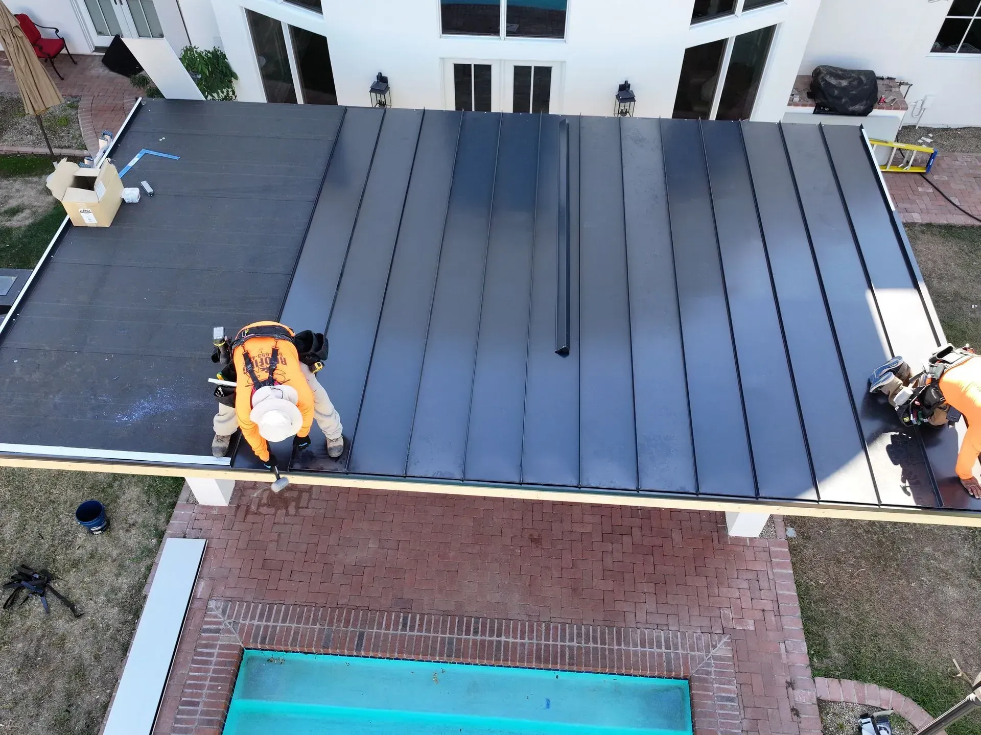 Workers installing dark roof panels on a flat rooftop beside a building and pool